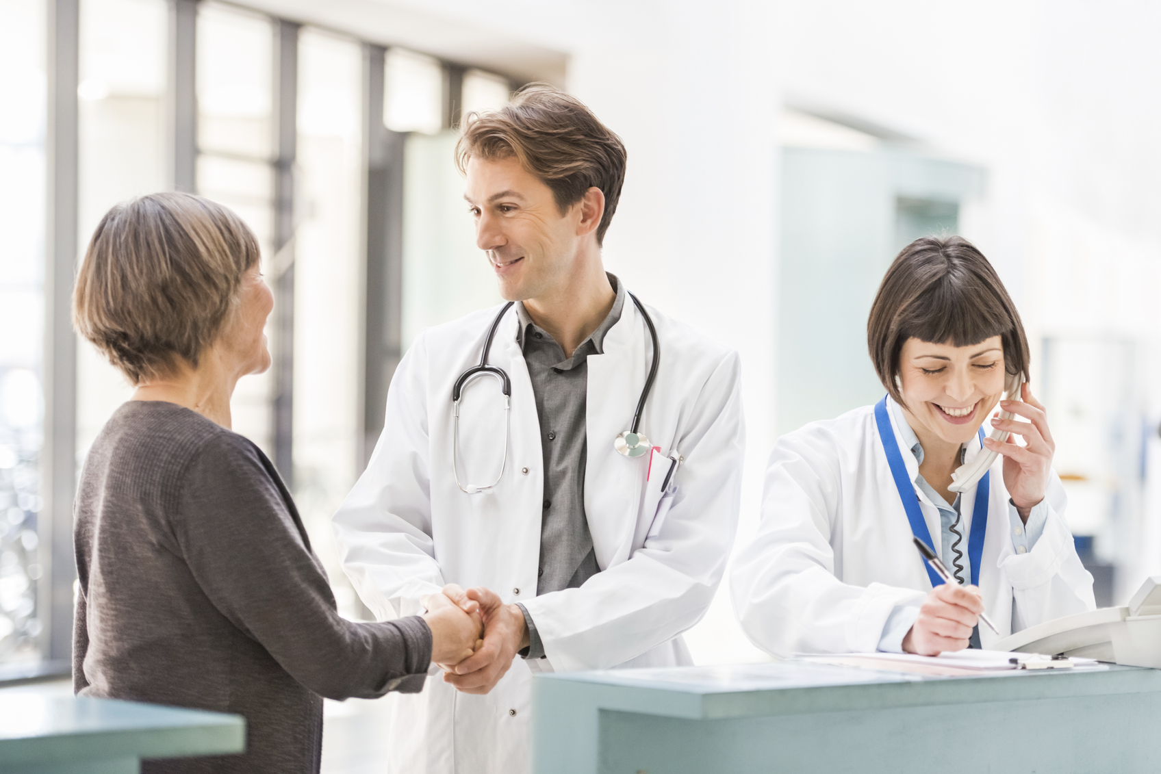 Doctor welcomes a senior patient at reception desk, while a colleague is calling on the phone.