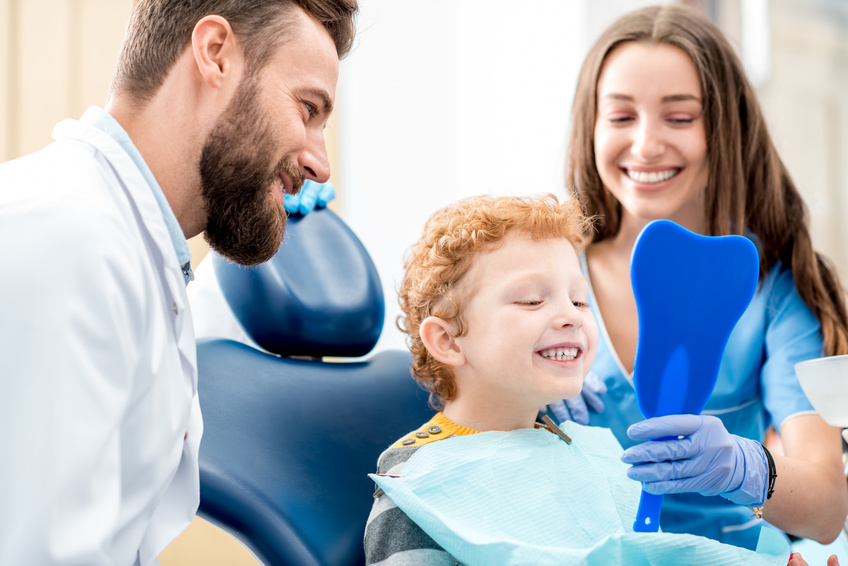 Boy with dentist at the dental office Young boy looking at the mirror with toothy smile sitting on the chair with dentist and assistant at the dental office